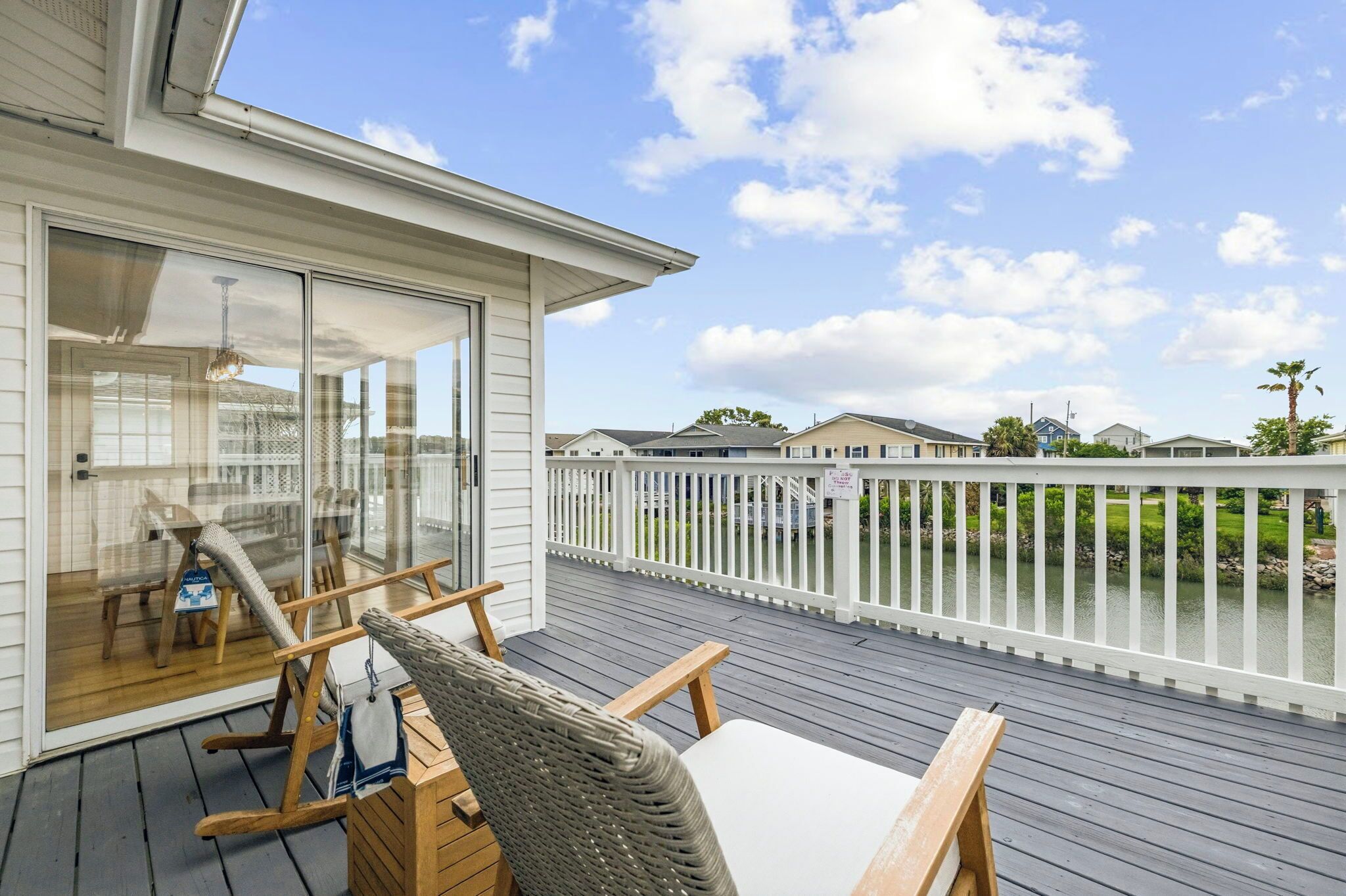 Um deck de madeira com mobiliário externo, uma porta de vidro que dá para uma área coberta e uma cerca branca com vista para a água.