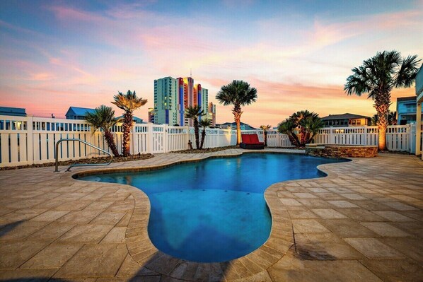 A swimming pool with a white fence, palm trees, and a cityscape in the background.