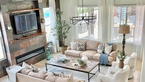 Ein modernes Wohnzimmer mit Kamin aus Stein, einem Fernseher, einer Kristallleuchter und einem großen Fenster mit Jalousien.