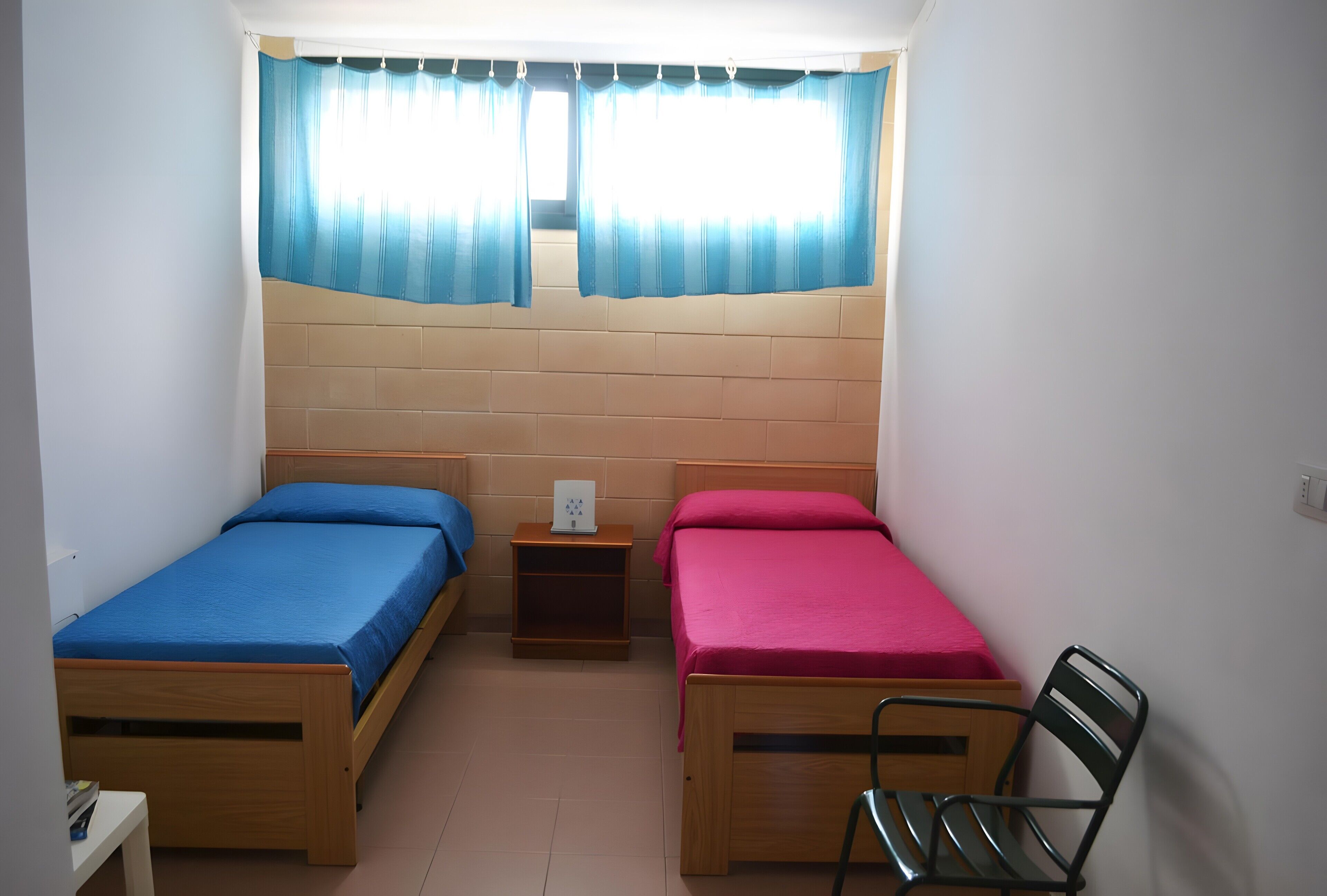 A dormitory room with two single beds, a small nightstand, a chair, and a window with curtains.