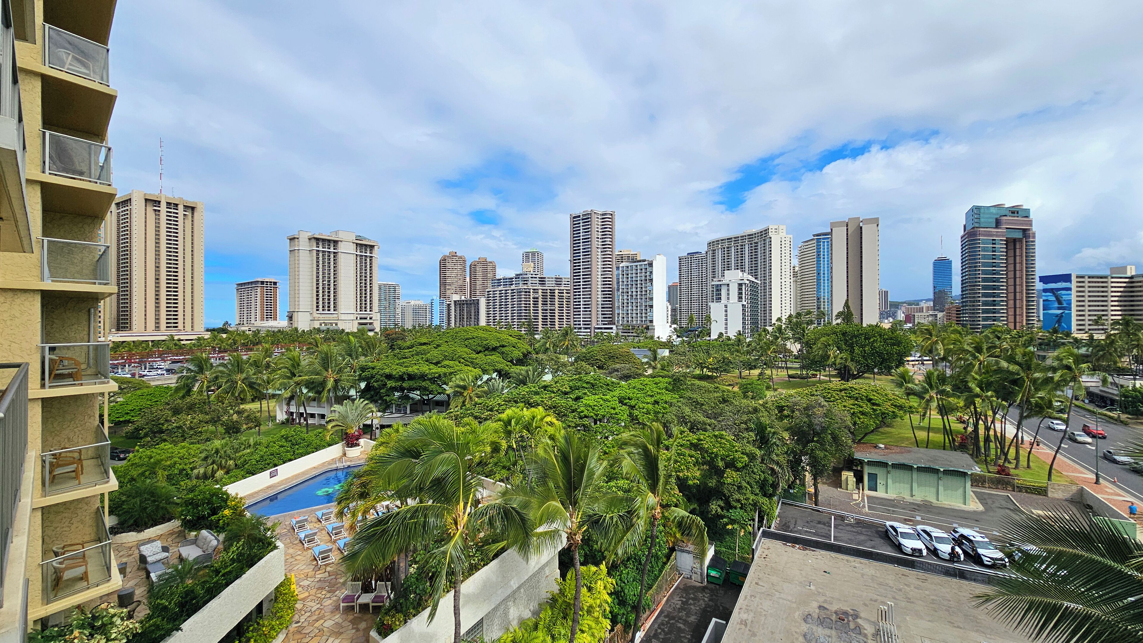 A high-rise building with a pool and outdoor seating area, surrounded by lush greenery and a city skyline in the background.