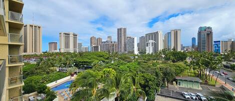 A high-rise building with a pool and outdoor seating area, surrounded by lush greenery and a city skyline in the background.