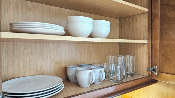 A wooden cabinet with shelves holding white plates, bowls, mugs, and clear glasses. - LUANA WAIKIKI HOTEL 502 🏝 2 Queen Bed CityViewStudio,WiFi,AC,GYM,POOL & Kitchen (Honolulu)