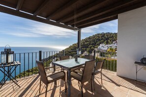 A terrace with a table and chairs, overlooking a coastal view with houses and greenery.