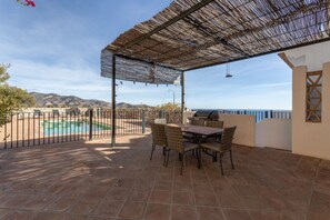 An outdoor patio with a table and chairs, a grill, and a view of mountains and the sea.
