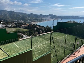 Un court de tennis offrant une vue sur une ville côtière et, au loin, sur des montagnes.