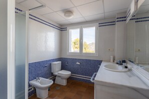 Une salle de bain comprenant des toilettes, un bidet et un lavabo, carrelée de blanc et de bleu, avec une fenêtre et un miroir.