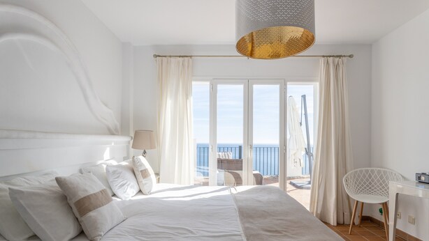 A bedroom with a bed, a desk, a chair, and a view of the sea.