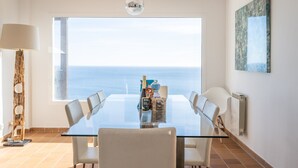 A dining area with a glass table, white chairs, a view of the sea, a hanging light fixture, and a wall-mounted artwork.