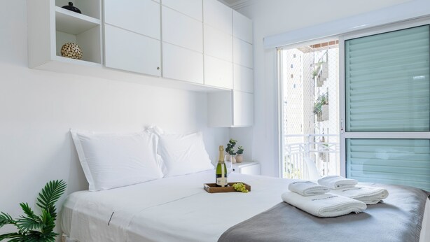 A neatly made bed with white linens, a bedside table with a bottle and grapes, and a potted plant. - AC, vaga, academia Vila Olímpia | Brazilian Corner (Sao Paulo)