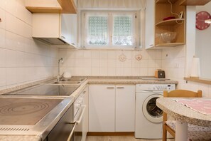 Une petite cuisine équipée d’une cuisinière, d’un évier et d’un lave-linge, une fenêtre ornée de rideaux en dentelle et une horloge au mur.