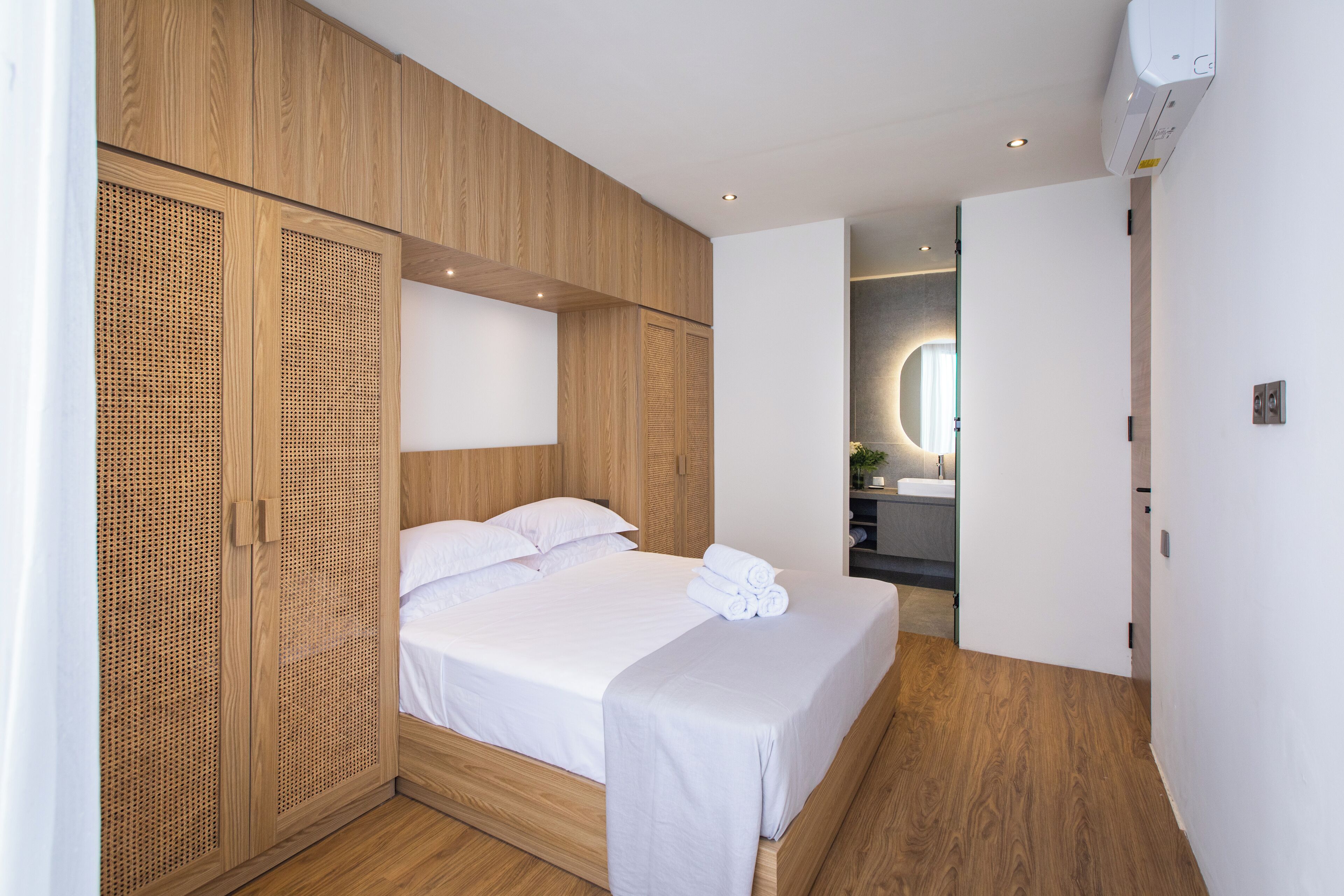 Une chambre moderne avec une armoire en bois, un lit recouvert de linge de maison blanc et une salle de bain visible à travers une porte ouverte.