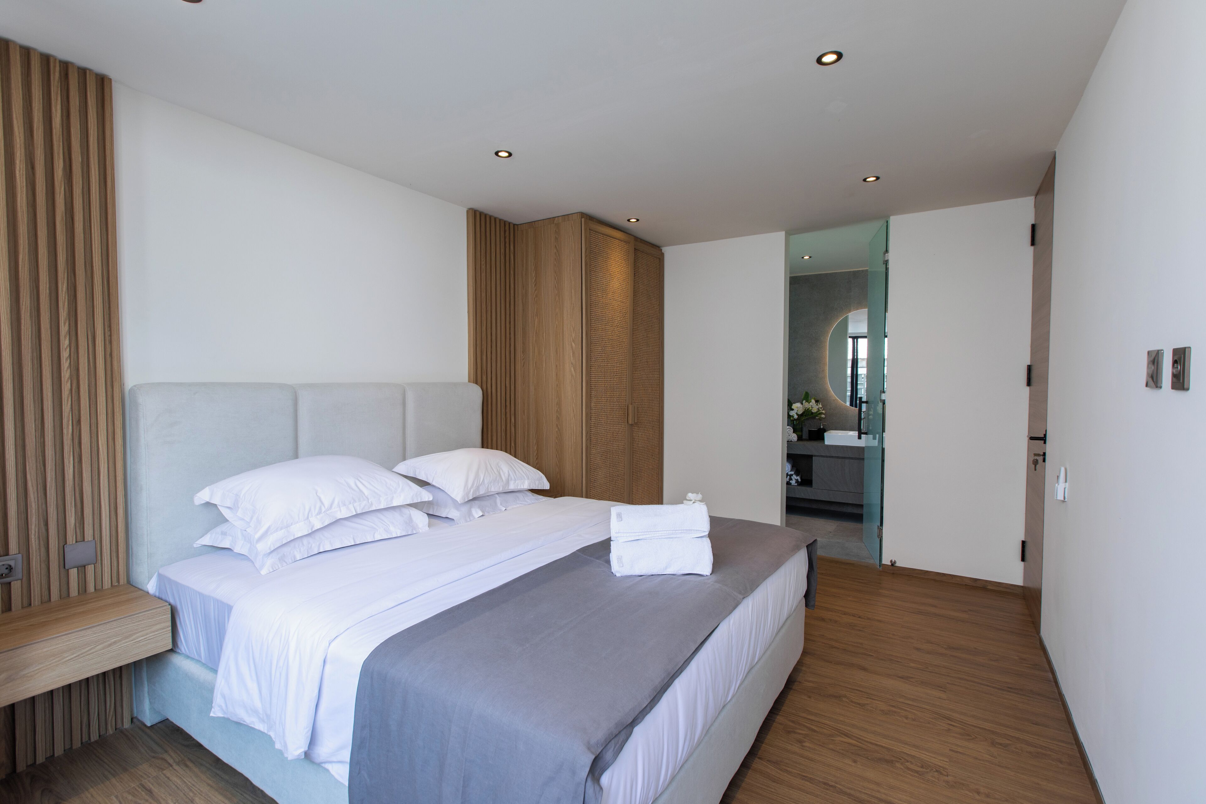 A modern bedroom with a large bed, wooden panel wall, and a view into the bathroom.