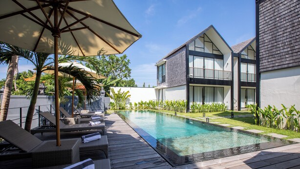 A modern pool area with lounge chairs and umbrellas, surrounded by a wooden deck and lush greenery.