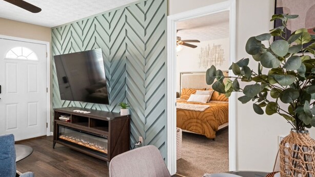 Ein Wohnzimmer mit Fernseher, einer Couch, einem Sessel, einer Wanddekoration und einem Durchgang zu einem Schlafzimmer.