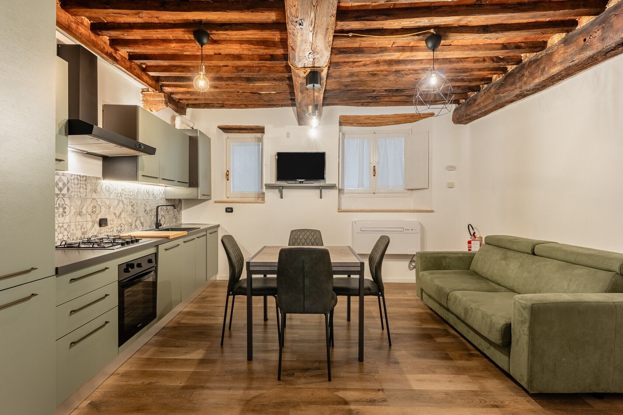 A kitchen with a dining table, chairs, a green sofa, and a TV mounted on the wall.