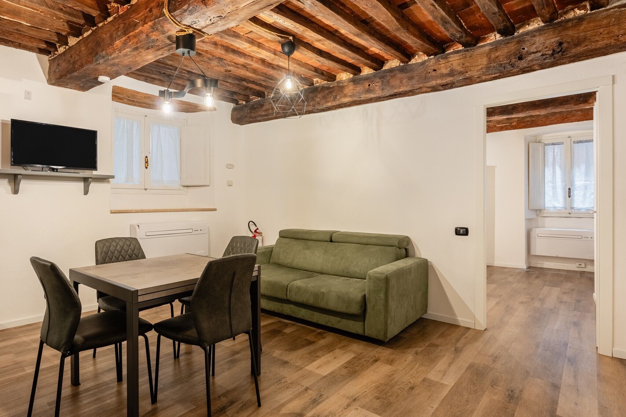 Une pièce avec un plafond en bois, un canapé vert, une table à manger avec des chaises, un téléviseur fixé au mur et une fenêtre avec des rideaux.