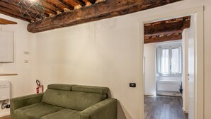 A room with a green sofa, wooden beams on the ceiling, a white wall, and a door leading to a bathroom.