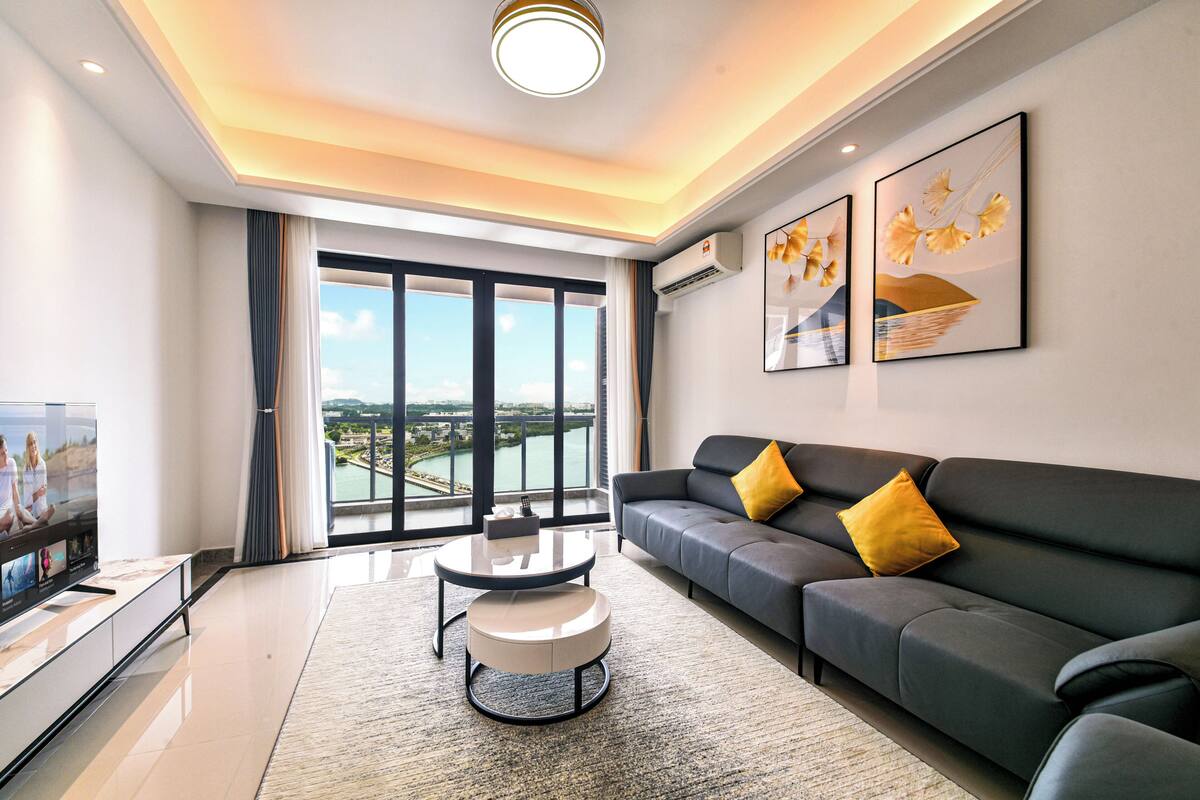 A modern living room with a large window, a sofa, a coffee table, and a flat-screen TV.