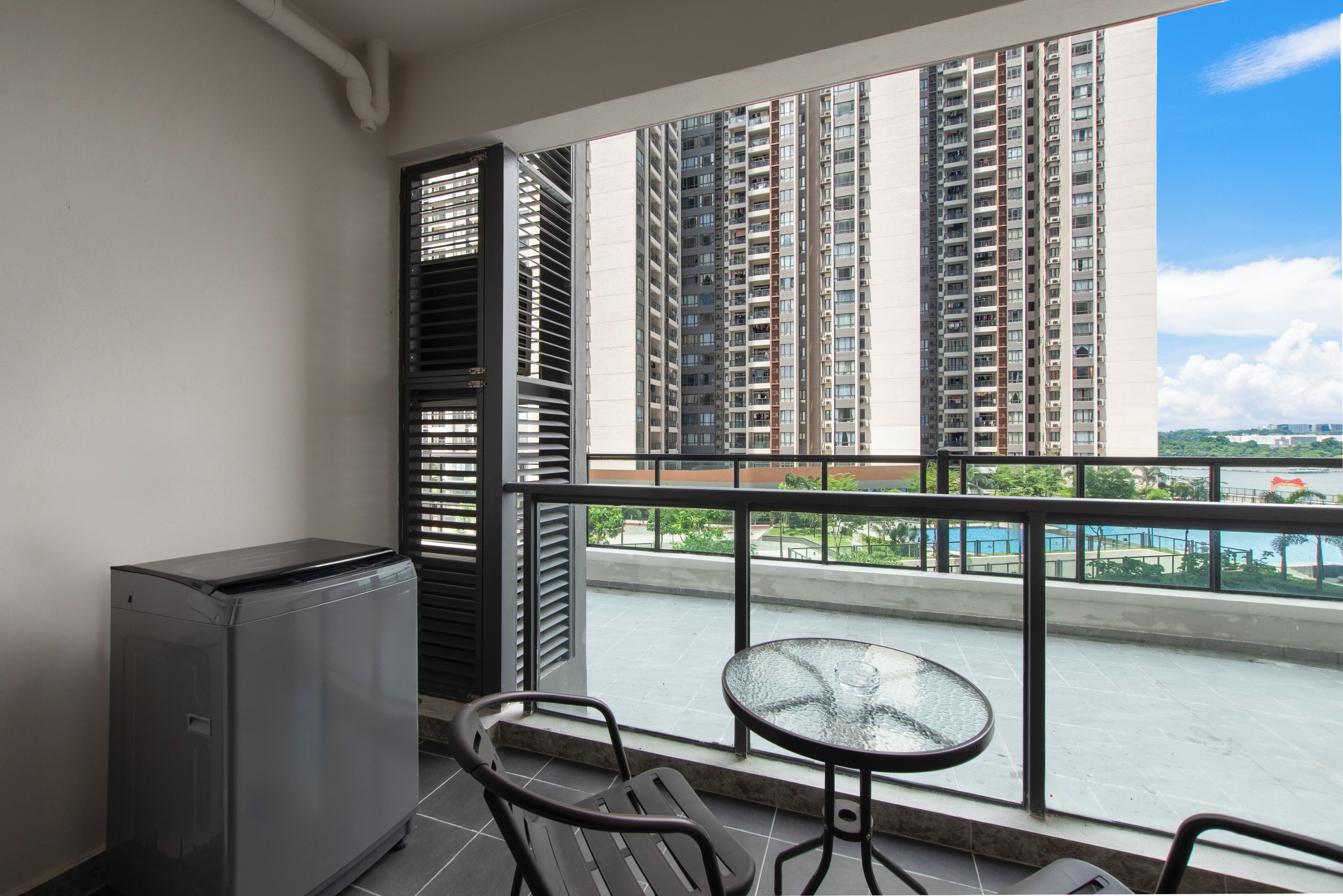 A balcony with a compact table and chairs, a small refrigerator, and a view of tall residential buildings and a swimming pool.