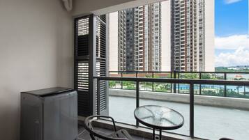 A balcony with a compact table and chairs, a small refrigerator, and a view of tall residential buildings and a swimming pool.