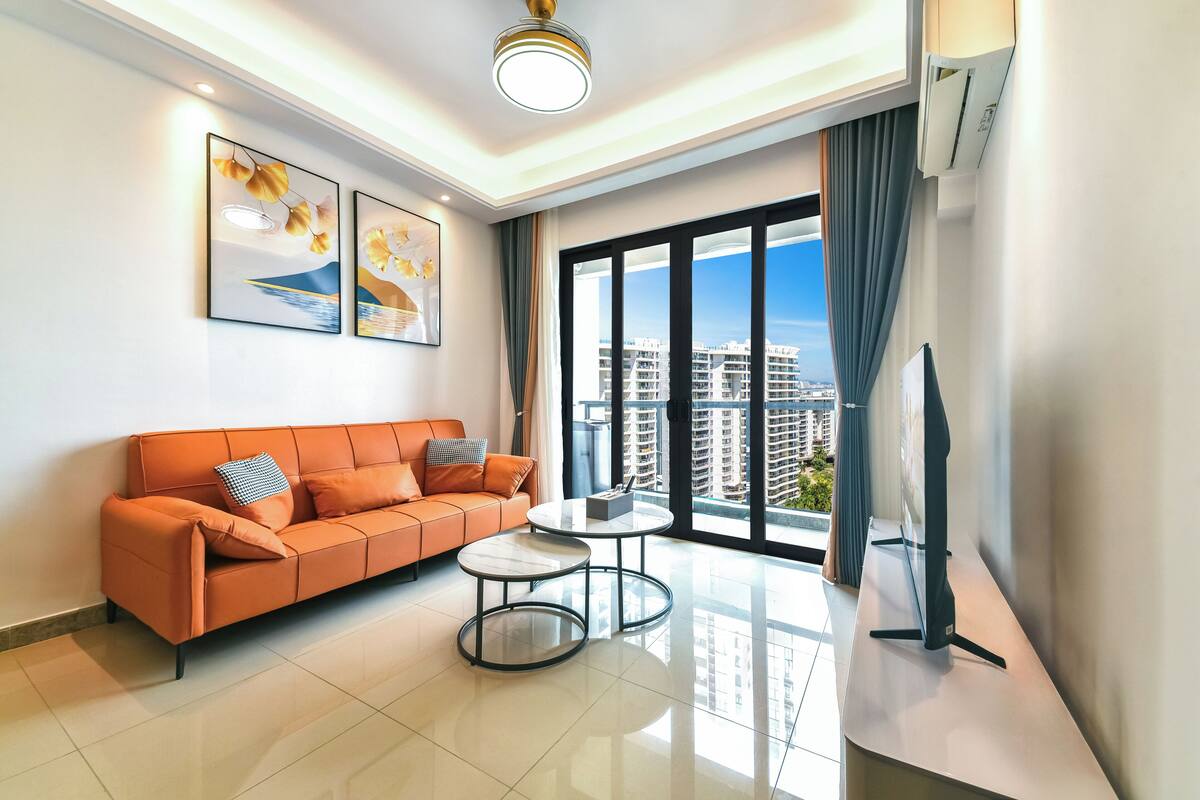 A modern living room with a sofa, two small tables, a flat-screen TV, and large windows with a city view.