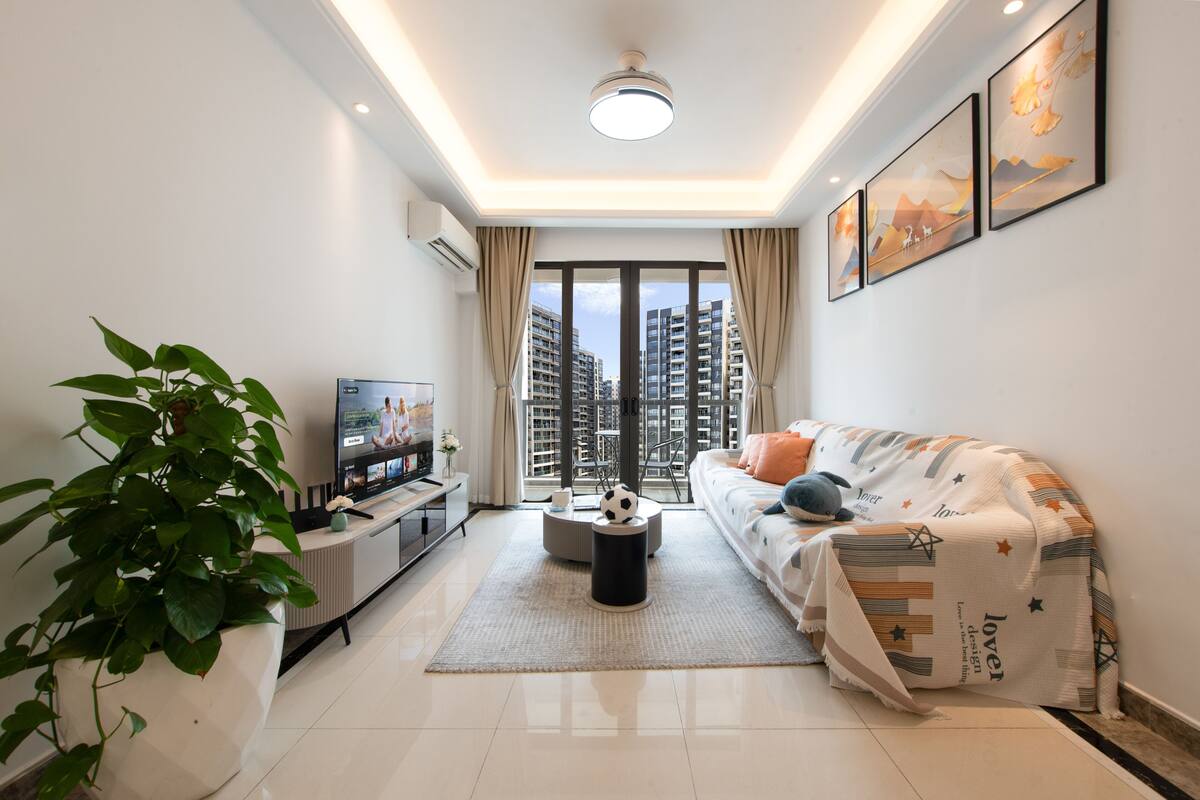 A modern living room with a sofa, TV, and a large window with a view of high-rise buildings.