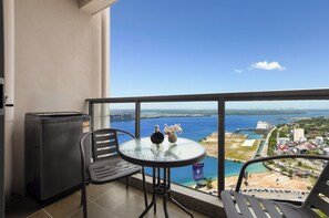A balcony with a glass table, two chairs, a small black appliance, and a view of a coastal cityscape.