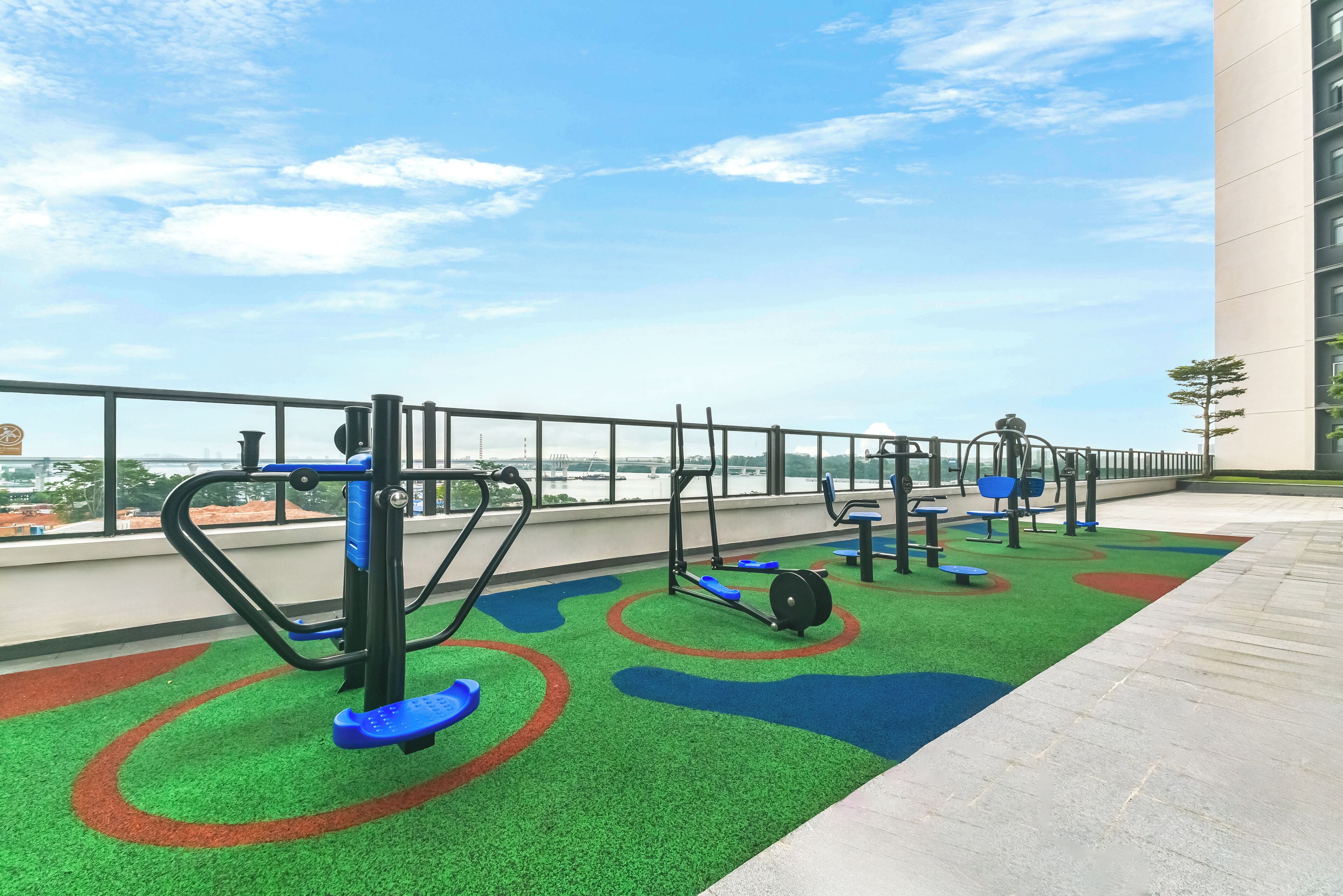 An outdoor gym area with exercise equipment, green artificial turf, and a view of a bridge.