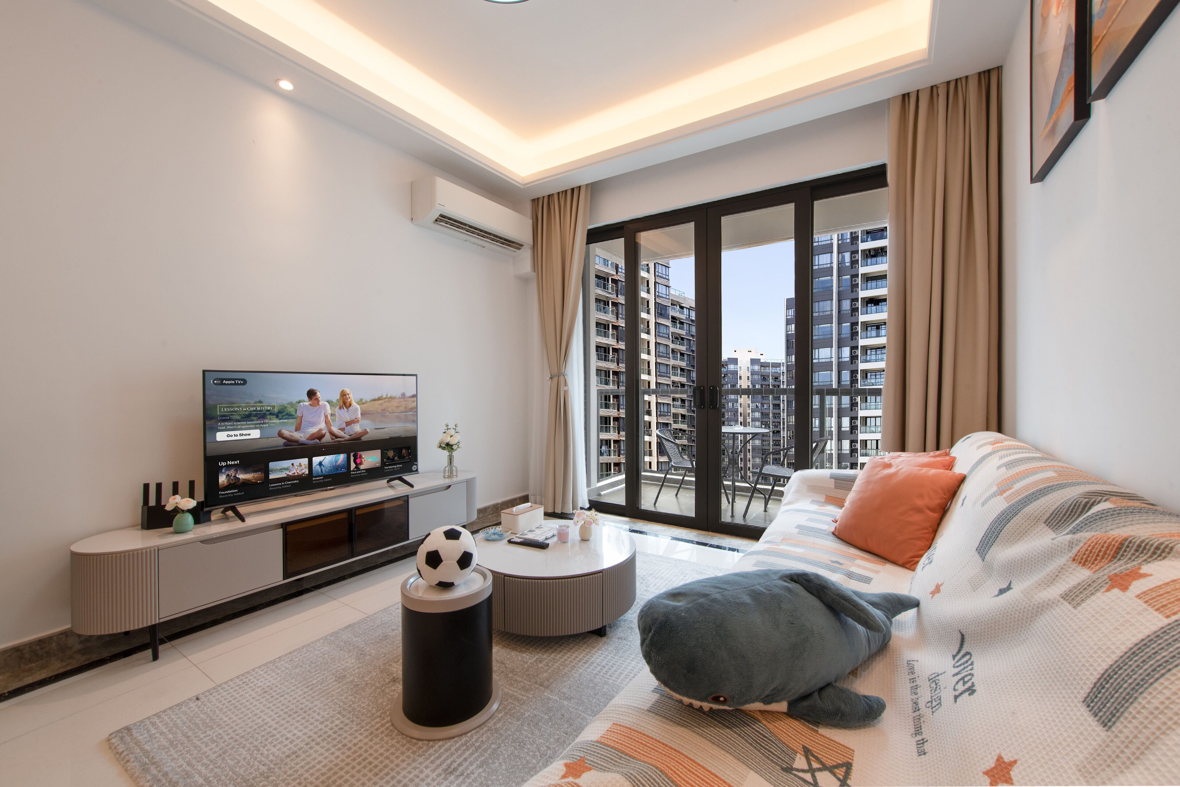 A modern living room with a flat-screen TV, a sofa, a coffee table, and a large window with a city view.
