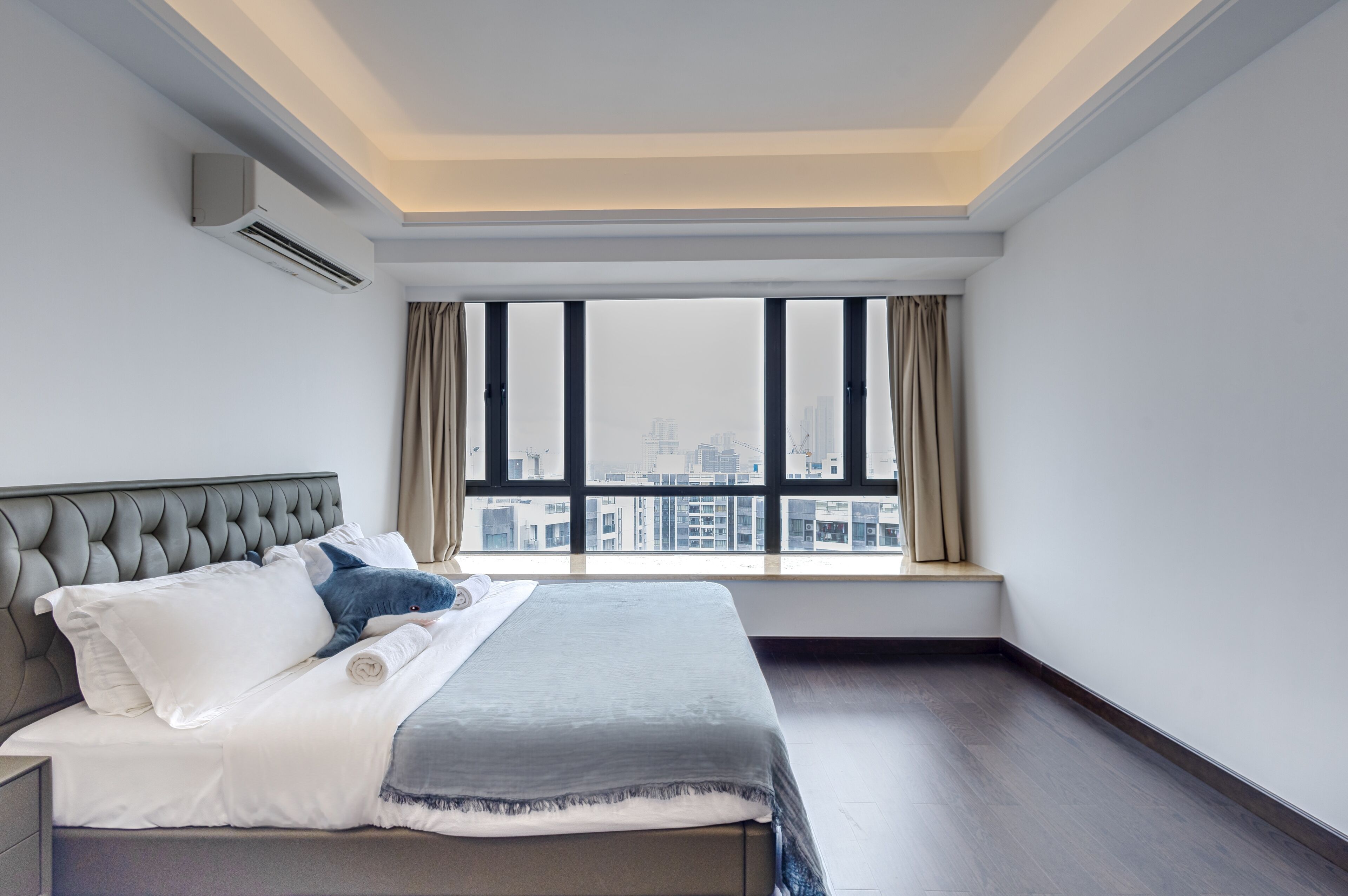 A modern hotel room with a large bed, a view of the city through floor-to-ceiling windows, and a built-in air conditioning unit.