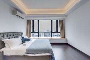 A modern hotel room with a large bed, a view of the city through floor-to-ceiling windows, and a built-in air conditioning unit.