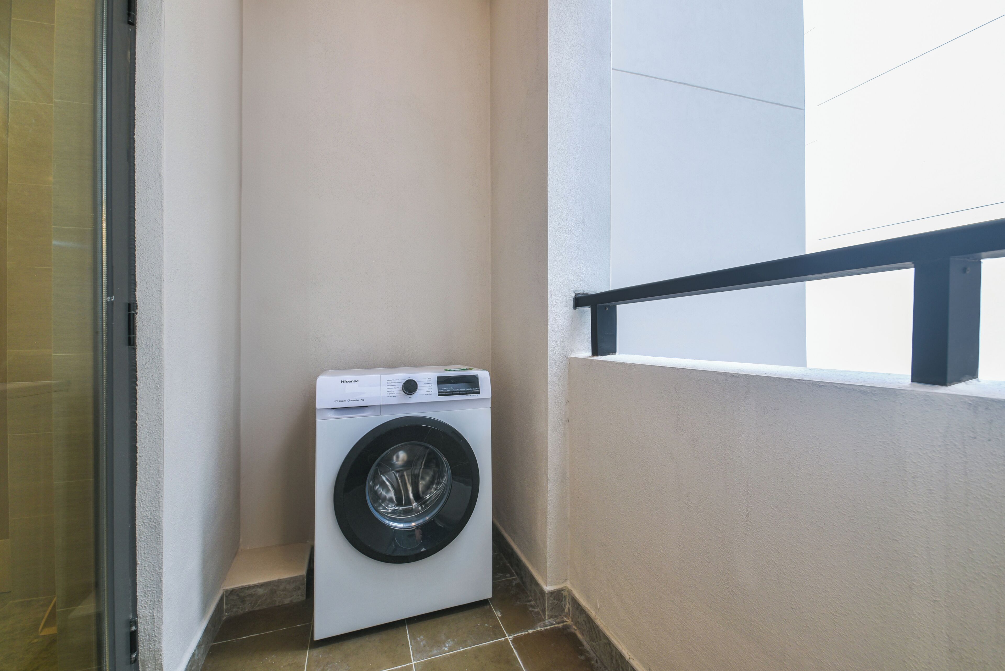 Une petite buanderie moderne, dotée d’un lave-linge à chargement frontal, d’une porte en verre et d’un balcon avec une rambarde noire.
