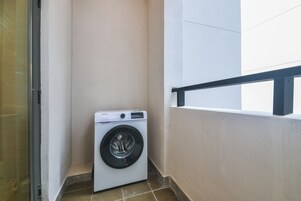 Une petite buanderie moderne, dotée d’un lave-linge à chargement frontal, d’une porte en verre et d’un balcon avec une rambarde noire.