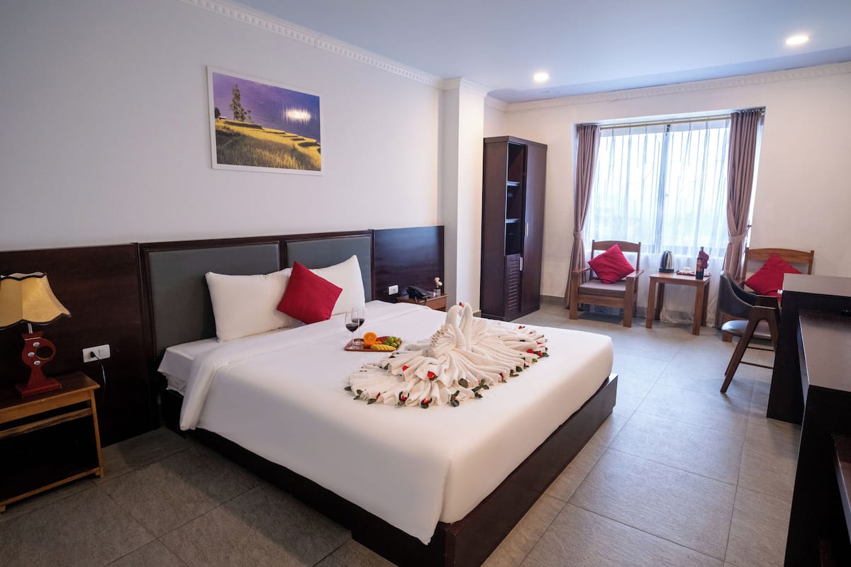 Habitación de hotel con una cama bien hecha, una mesita de noche con lámpara, un escritorio con silla y una ventana con cortinas.