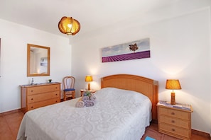 Quarto bem arrumado com cama, mesas de cabeceira, cômoda, cadeira e um quadro na parede.