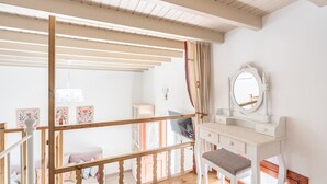 Une pièce avec un plafond en bois, une coiffeuse blanche avec un miroir, un banc et un lit avec une literie aux couleurs vives.