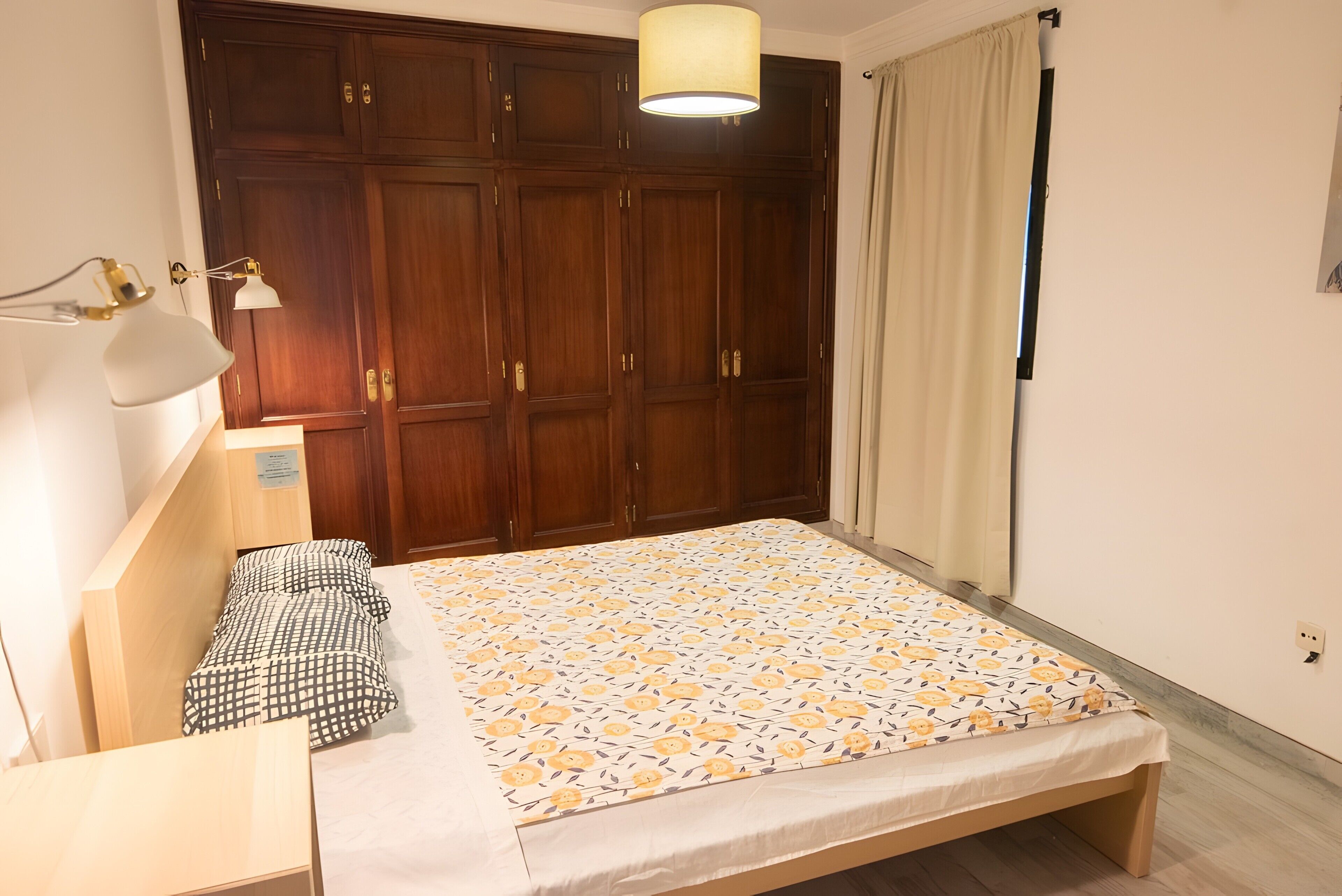 Une chambre à coucher comprenant un lit, une table de chevet, une lampe, une armoire et une fenêtre avec des rideaux.