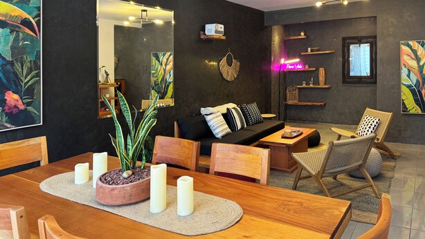 A modern living room with a wooden dining table, chairs, a sofa, and a coffee table. There are decorative plants, a mirror, and artwork on the walls.