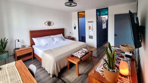 A modern bedroom with a wooden bed, bedside tables, a desk, a chair, a potted plant, and a television.
