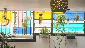 A room with a vibrant window, a hanging light fixture, a cushioned chair, and various potted plants.