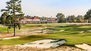 Een golfbaan met zandbunkers, bomen en gebouwen op de achtergrond.