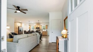 A modern kitchen with wooden cabinets, a dining area with a table and chairs, and a living room with a sofa and a ceiling fan. - The Porch and Pines Cottage (Moore County)