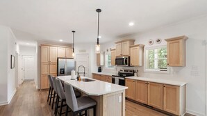 A modern kitchen with wooden cabinets, a central island with bar stools, stainless steel appliances, and pendant lighting. - The Porch and Pines Cottage (Moore County)
