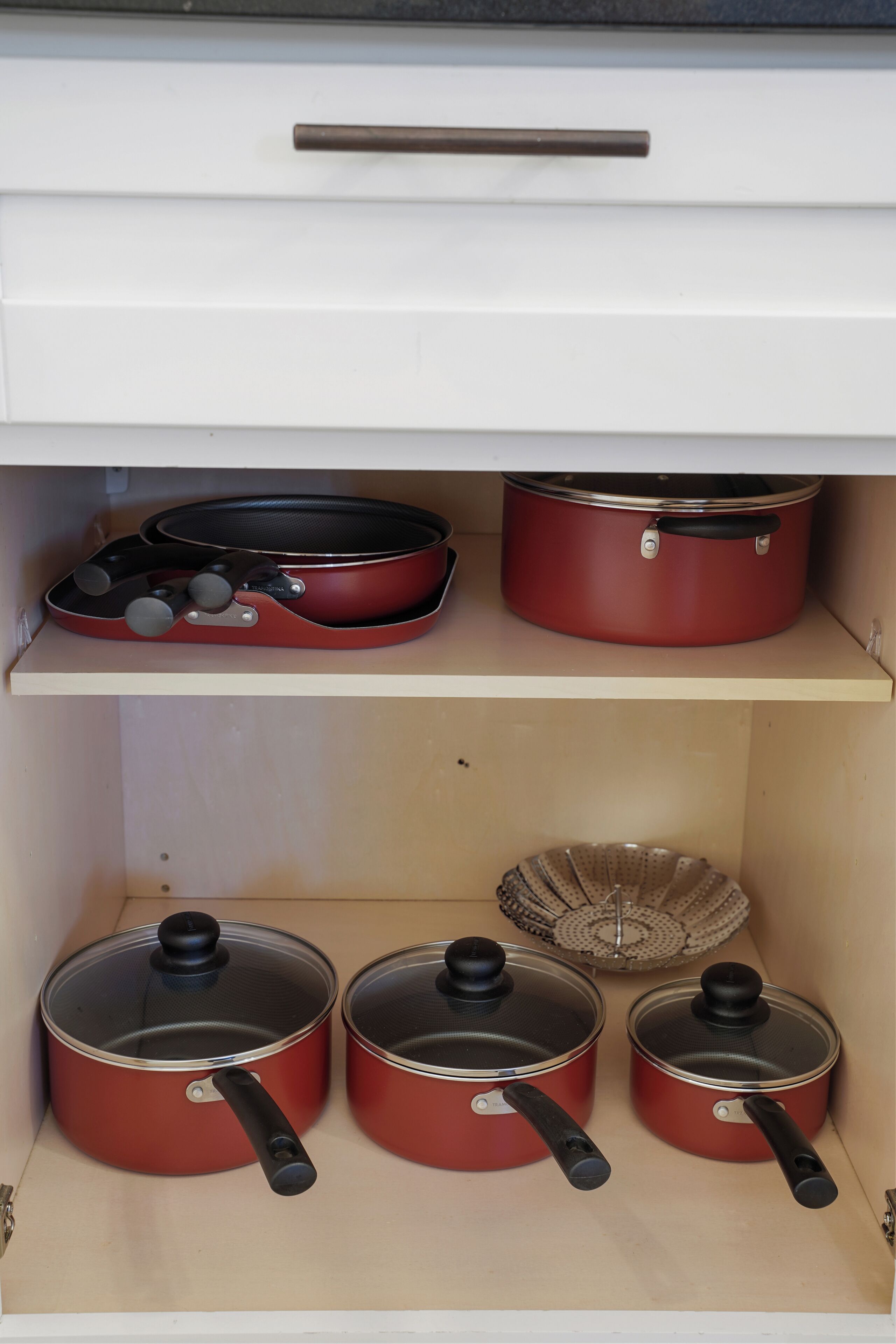 A kitchen cabinet with three red pots and pans, a frying pan, and a colander.