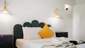Un lit bien fait, avec du linge de lit blanc, une tête de lit recouverte d’un tissu vert et un coussin jaune. Il y a deux tables de chevet, l’une avec une lampe et l’autre avec un téléphone. La pièce a des murs blancs et une porte visible au fond.