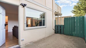 Un immeuble résidentiel doté d’une grande fenêtre, d’une porte, d’une terrasse en béton et d’une clôture verte.