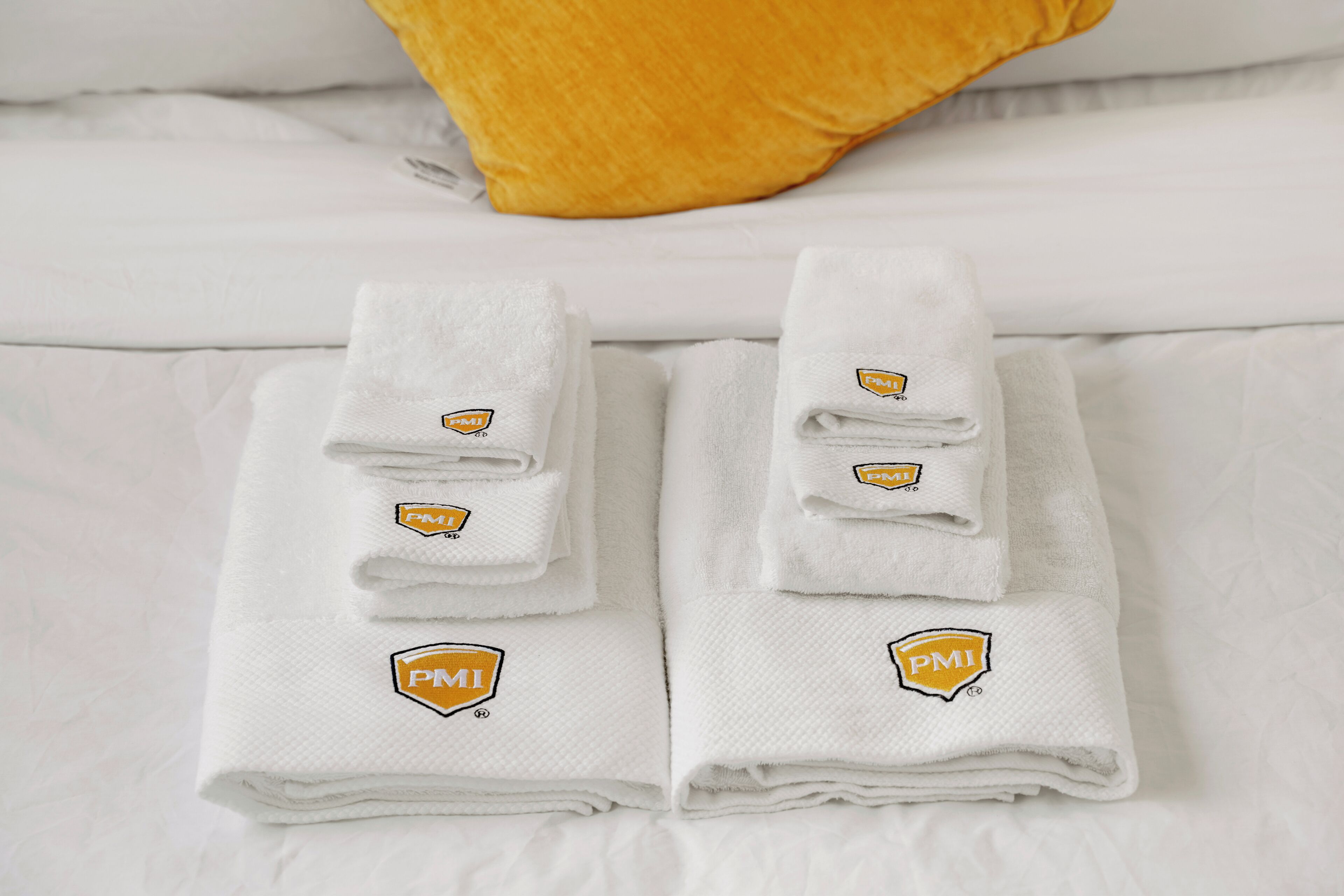 Two neatly folded white towels with a logo on a bed with white sheets and a yellow pillow.