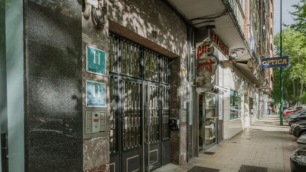 Une rue bordée d’un immeuble doté d’une grille en métal, d’une enseigne indiquant « Optica » et de voitures garées.