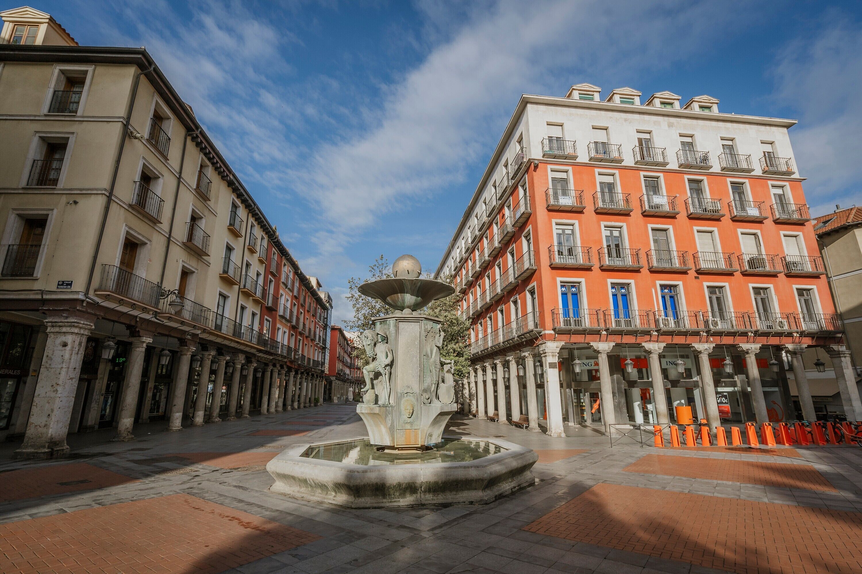 Une place publique avec une fontaine centrale, des bâtiments dotés de balcons et un ciel dégagé.